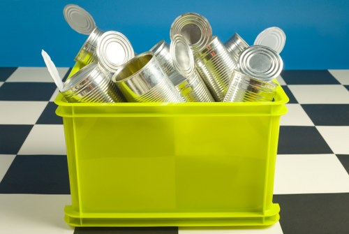A bright yellow plastic bin filled with topped and leaning silver aluminium cans, some with visible ridges and open ends, placed on a black and white checkered floor against a blue wall background. The cans are stacked haphazardly within the container, which appears to be used for waste collection or recycling purposes. The environment suggests an indoor setting, possibly part of a waste removal or recycling service involved with rubbish disposal in Hornsey, as indicated by the website context. The clear visual focus is on the collection of metal cans, with the colours and textures providing clarity on the materials. The image's lighting is even, highlighting the metallic sheen of the cans and the vibrant colour of the bin, reinforcing the theme of waste management in a professional, straightforward manner related to rubbish removal services offered by Business Waste Removal Hornsey.