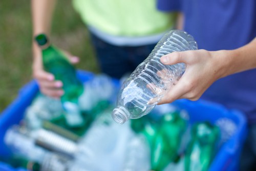 A close-up of a person's hand gripping a clear plastic water bottle, which has a ridged body and a slightly tapered neck. The individual appears to be in the process of discarding the bottle, with other discarded plastic bottles and waste materials visible in a blue recycling container behind them. The background shows the lower half of another person wearing a yellow top and a black wristwatch, suggesting an outdoor setting, possibly on a driveway or pavement near Hornsey. The scene captures a moment of waste disposal related to rubbish clearance, aligning with services offered by Business Waste Removal Hornsey focused on recycling and rubbish removal. The environment is well-lit, indicating daytime, with a focus on the plastic waste and the action of discarding it, emphasizing waste management and recycling efforts typical of a professional waste removal service in the local area.