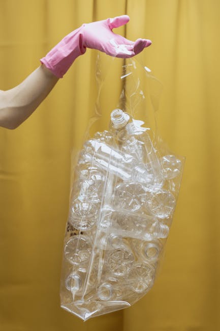 A person wearing a pink rubber glove is holding a large, clear plastic bag filled with empty transparent plastic bottles. The bottles vary in size and shape, some with ridged textures and others smoot
